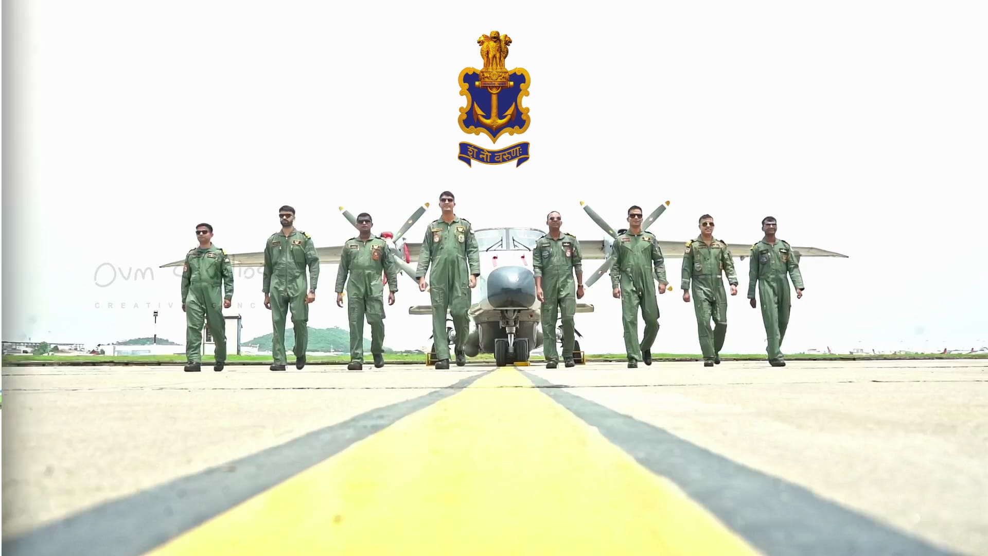 Indian Navy pilots in green flight suits walking in formation on an airstrip, with a propeller aircraft in the background and the Indian Navy insignia above them.